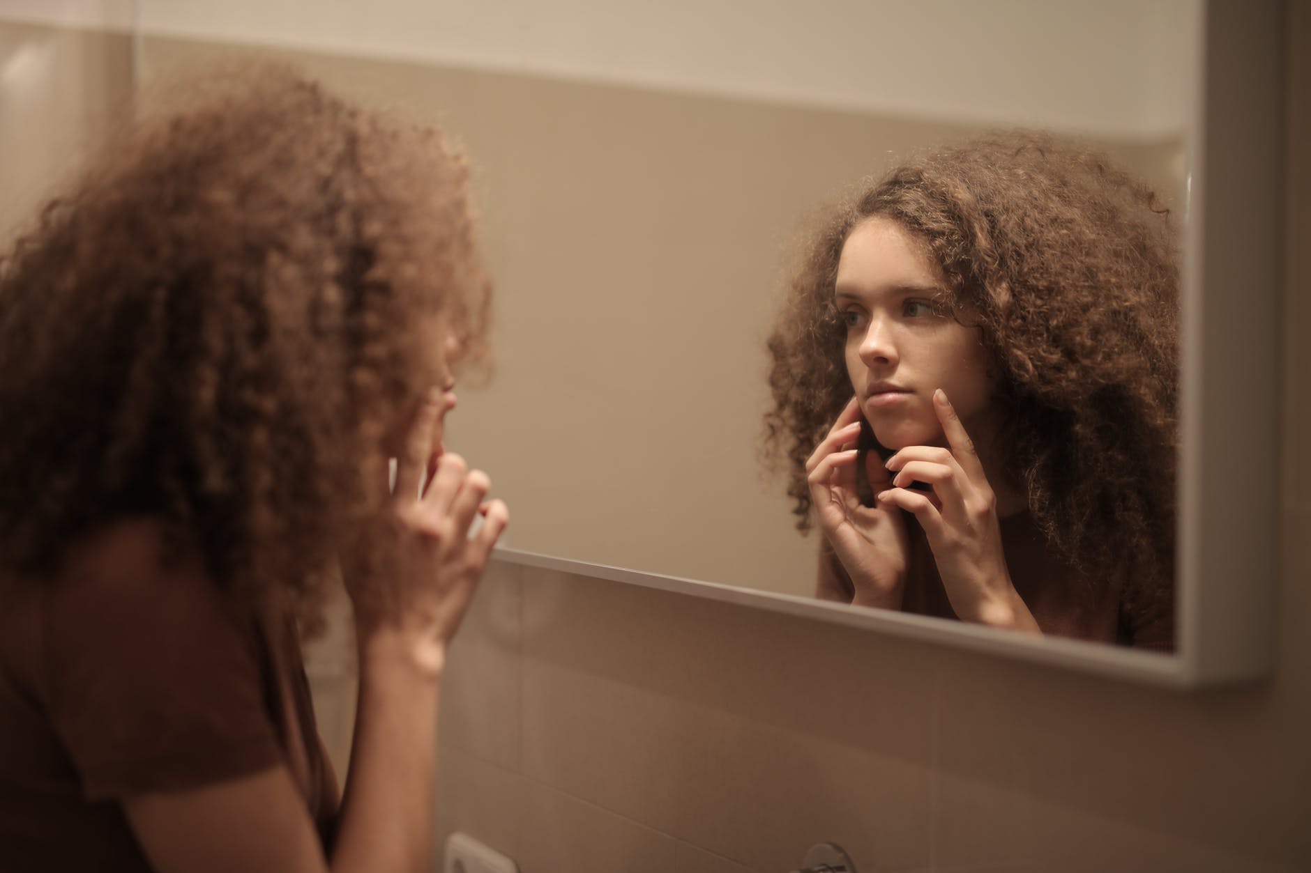 serious young lady touching face while looking at mirror in bathroom